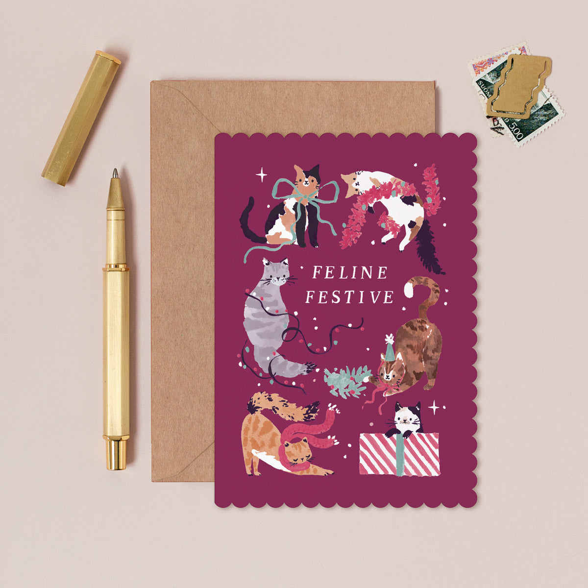Feline Cat Christmas card with cats in festive attire on a pink background, accompanied by a gold pen and brown envelope.