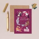 Feline Cat Christmas card with cats in festive attire on a pink background, accompanied by a gold pen and brown envelope.
