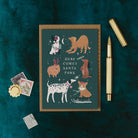 Christmas card with illustrations of dogs and text 'Here Comes Santa Paws' on a dark green background, with a pen and decorative items around.