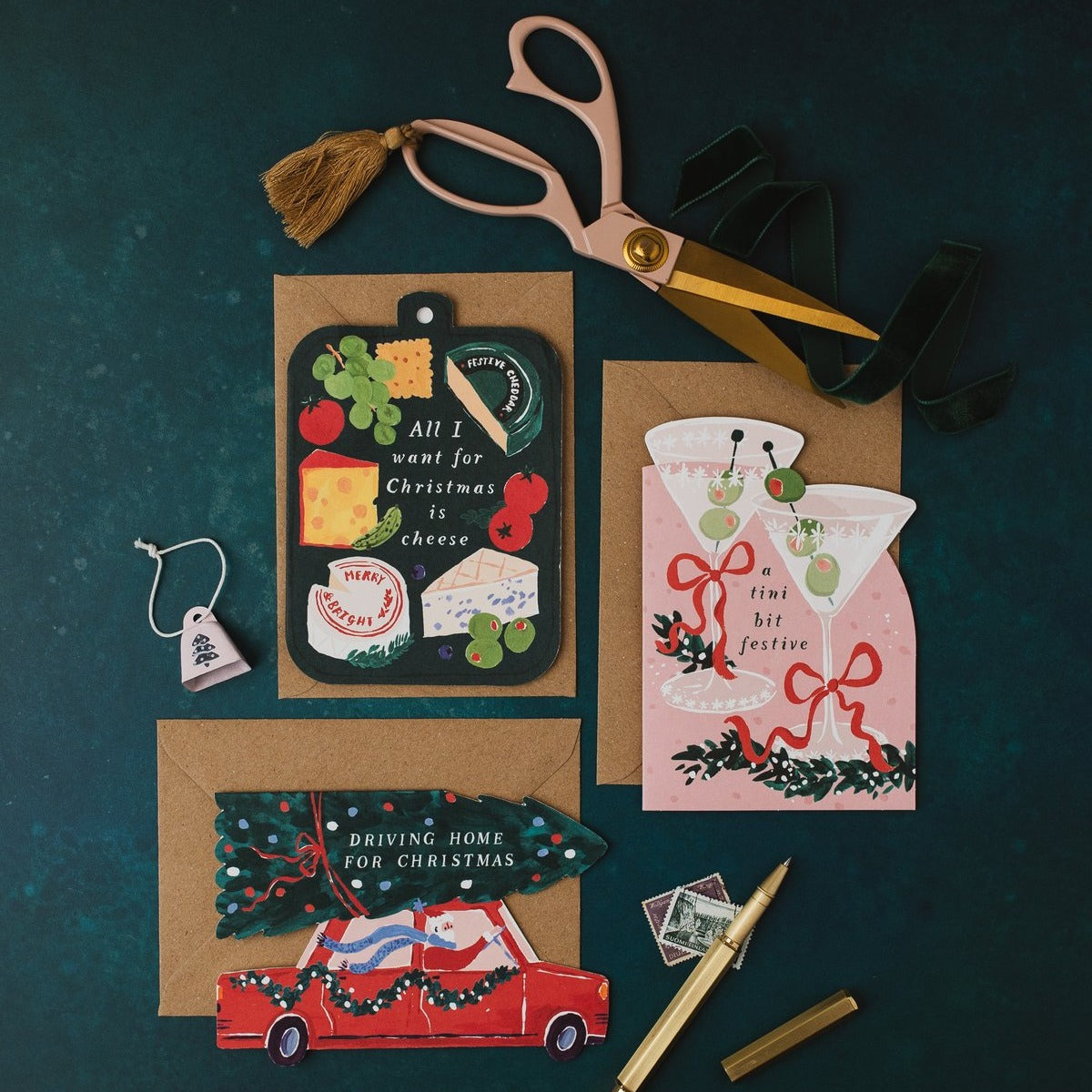 Cheeseboard, martini and Driving Home for Christmas shaped cards with kraft envelopes and gold scissors on a dark teal background