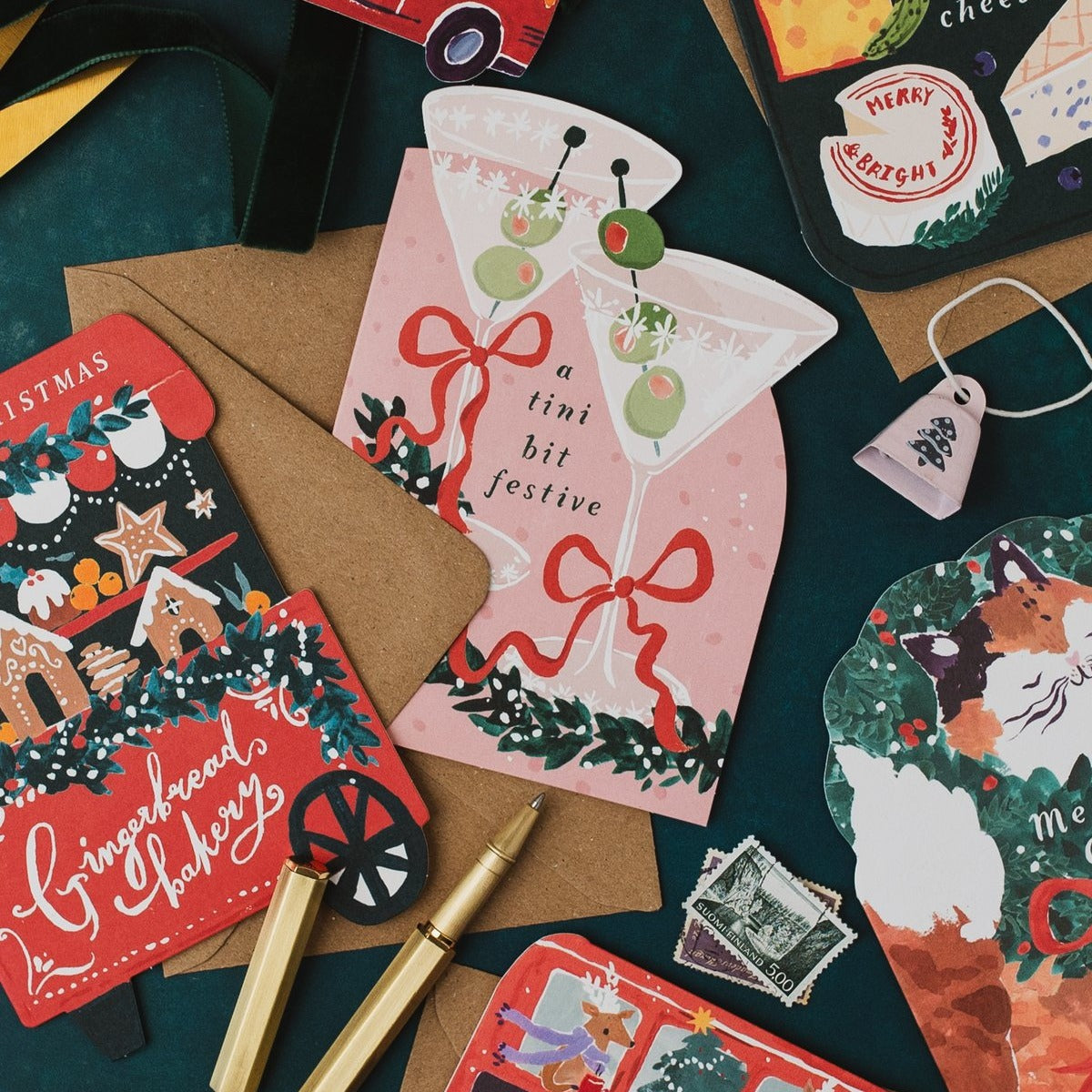 Gingerbread cart Christmas card with a gold pen on a dark teal background beside a martini Christmas card