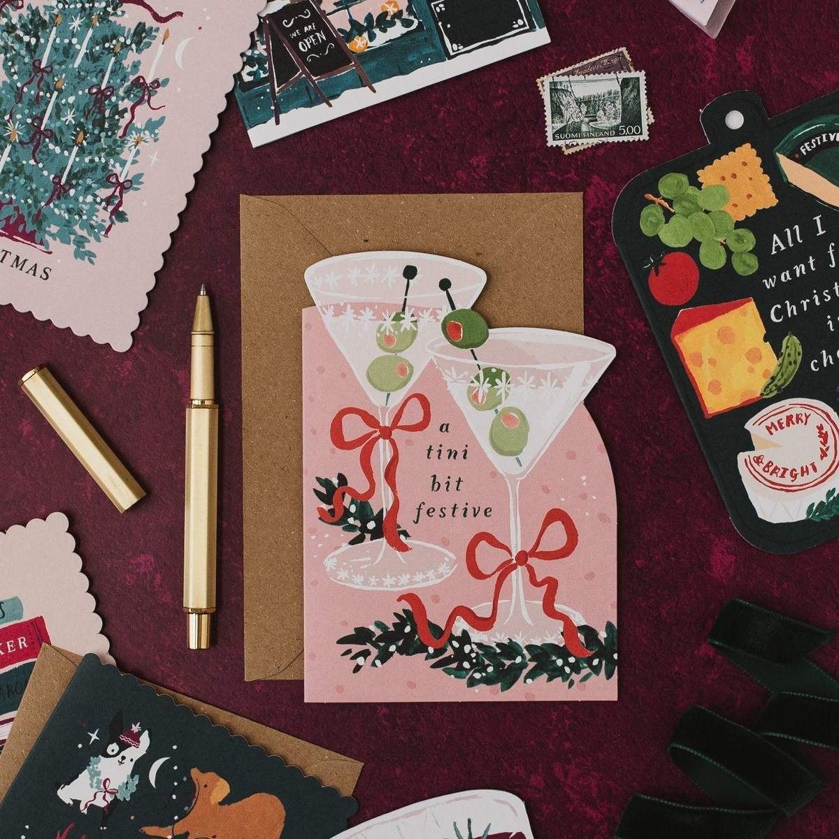 Tini Bit Festive martini shaped Christmas card with a gold pen on a deep red background, with Tree-mendous and cheeseboard Christmas cards